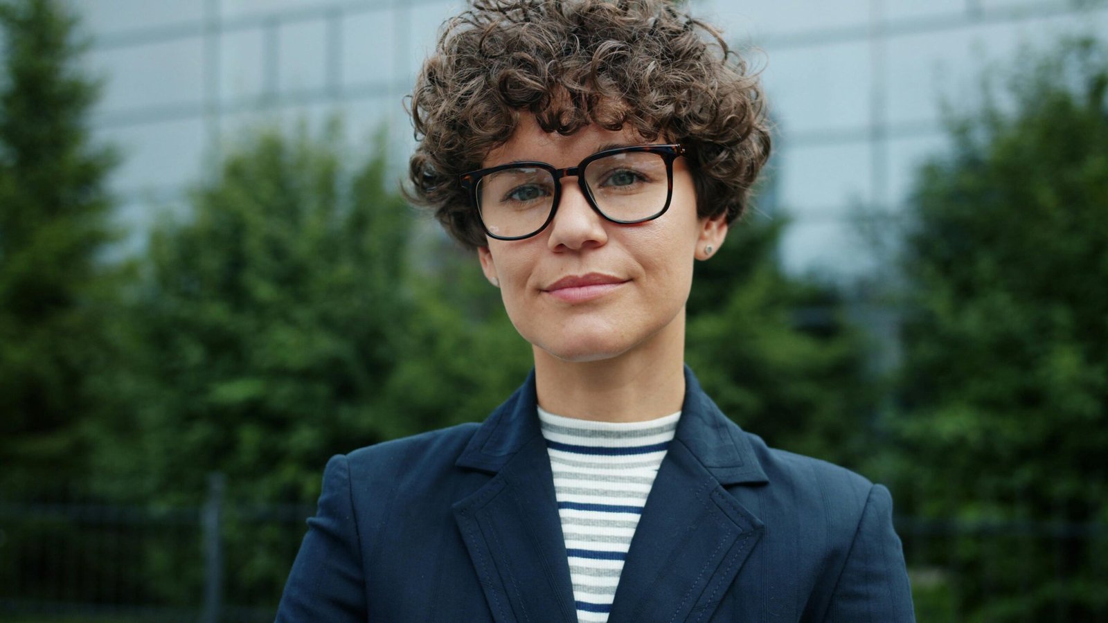 Portrait of a confident woman in business attire with curly hair and glasses outdoors.